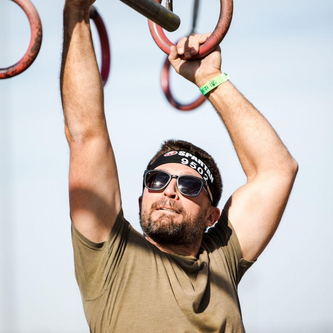 Man hanging from red rings with sunglasses and a headband against a light blue sky.